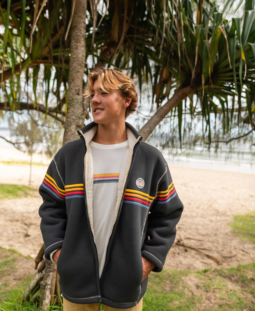 A young man stands outdoors by palm trees and the ocean, wearing a Golden Breed Reef Bonded Sherpa Jacket in Wash Black with colorful stripes over a white t-shirt.