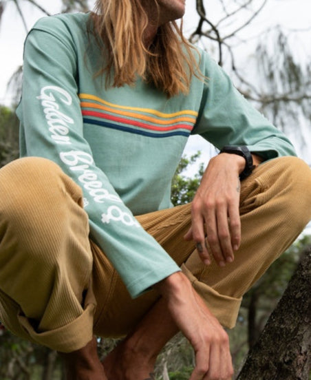 A person with long hair sits cross-legged outdoors, wearing a sage Heavy Island Long Sleeve Tee by Golden Breed with tan pants—embodying a relaxed beach surf lifestyle among trees and branches.