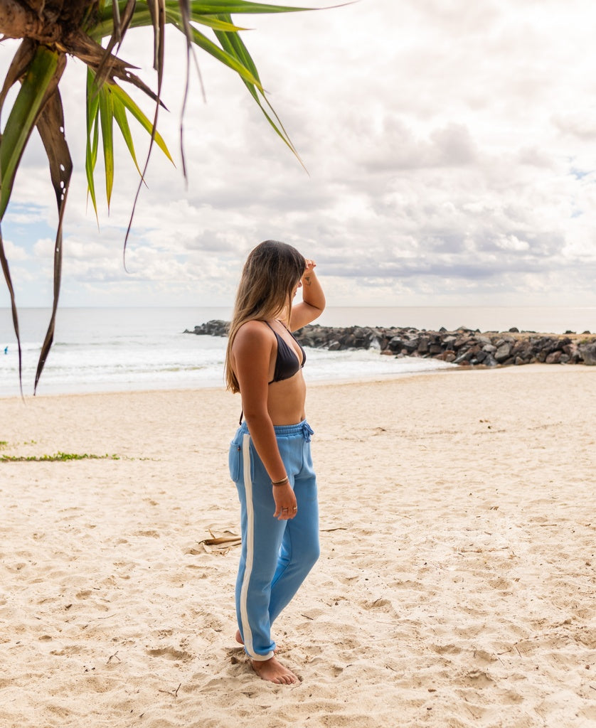 A woman wearing Golden Breed’s Retro Crunchie T/P trackpants in Azure/Off White stands barefoot on a sandy beach, gazing at the ocean under a cloudy sky.