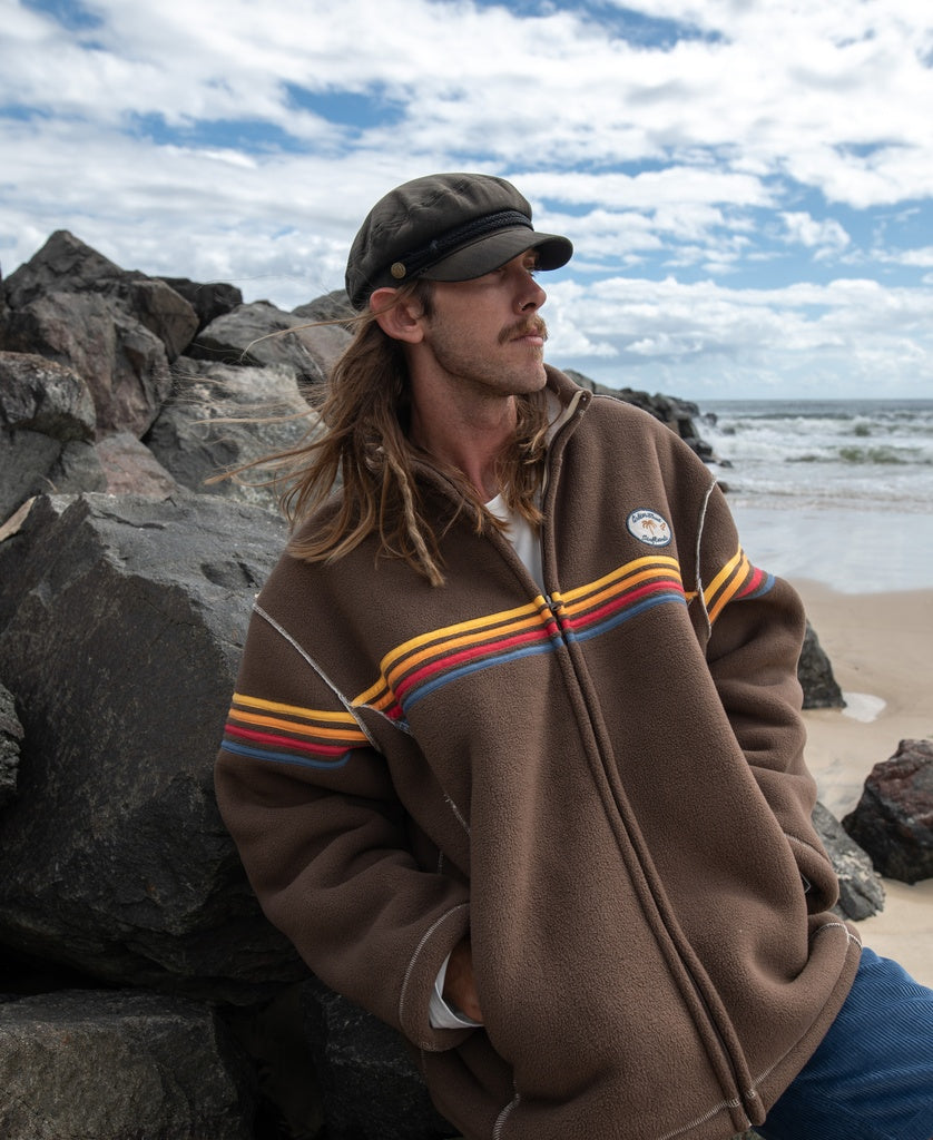 A man with long hair and a mustache, wearing the Golden Breed Reef Bonded Sherpa Jacket in Wash Brown and a black cap, stands by rocks on a beach under a partly cloudy sky.