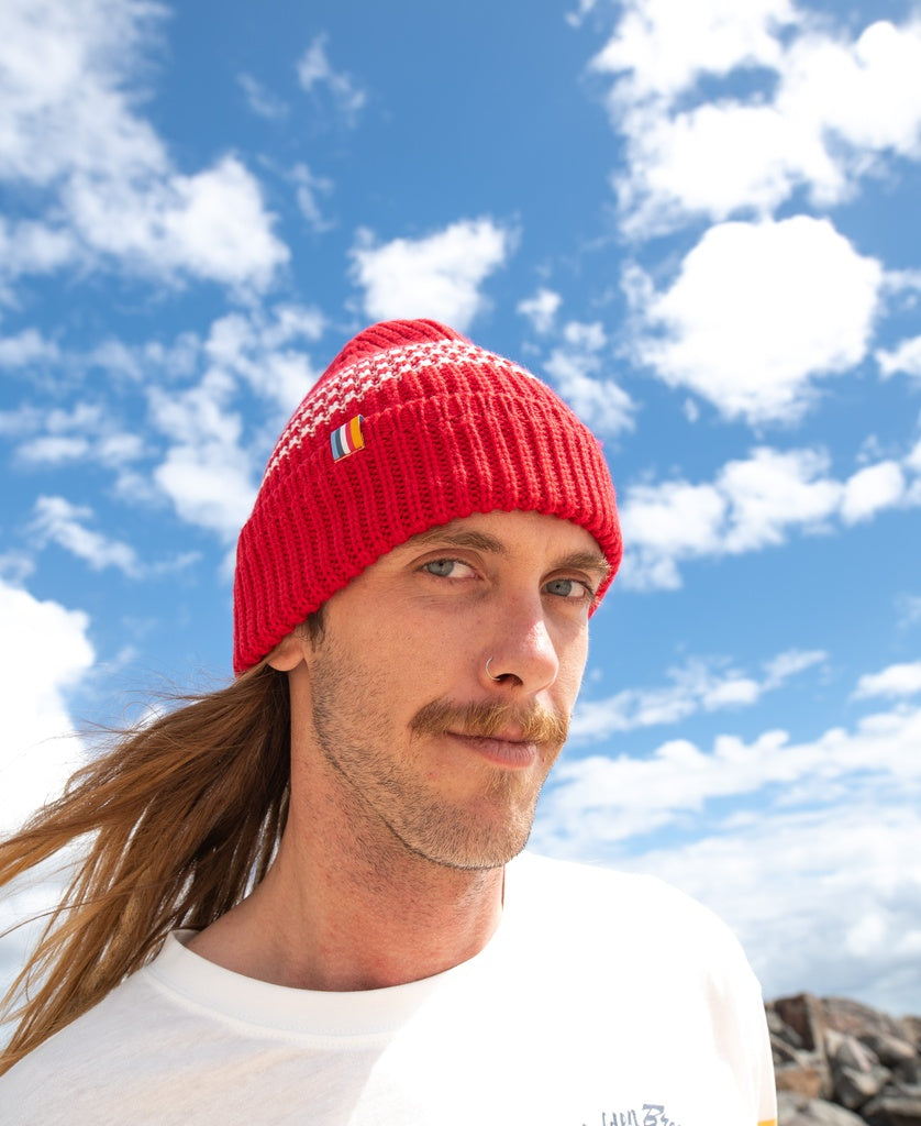 A man with long hair and a mustache wears the Golden Breed Beach Boy Wool Beanie in red and a white shirt, standing outdoors beneath a blue sky with clouds.