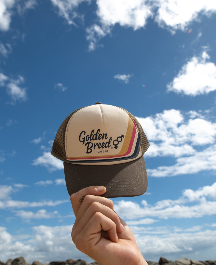 A hand holds up a Retro Trucker Cap | Wash Brown by Golden Breed, featuring front branding and an adjustable strap, set against a blue sky with scattered clouds.