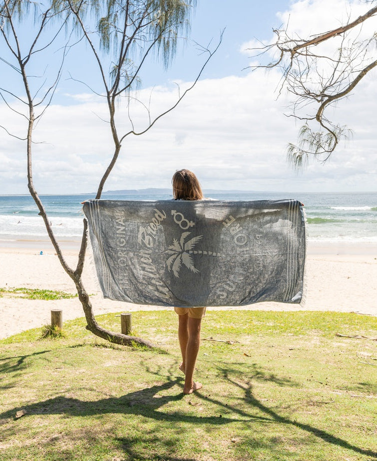A person stands on grass near the beach, holding the Golden Breed GB Otto Towel in Navy, a soft Turkish cotton towel featuring a palm tree design, while facing the ocean.