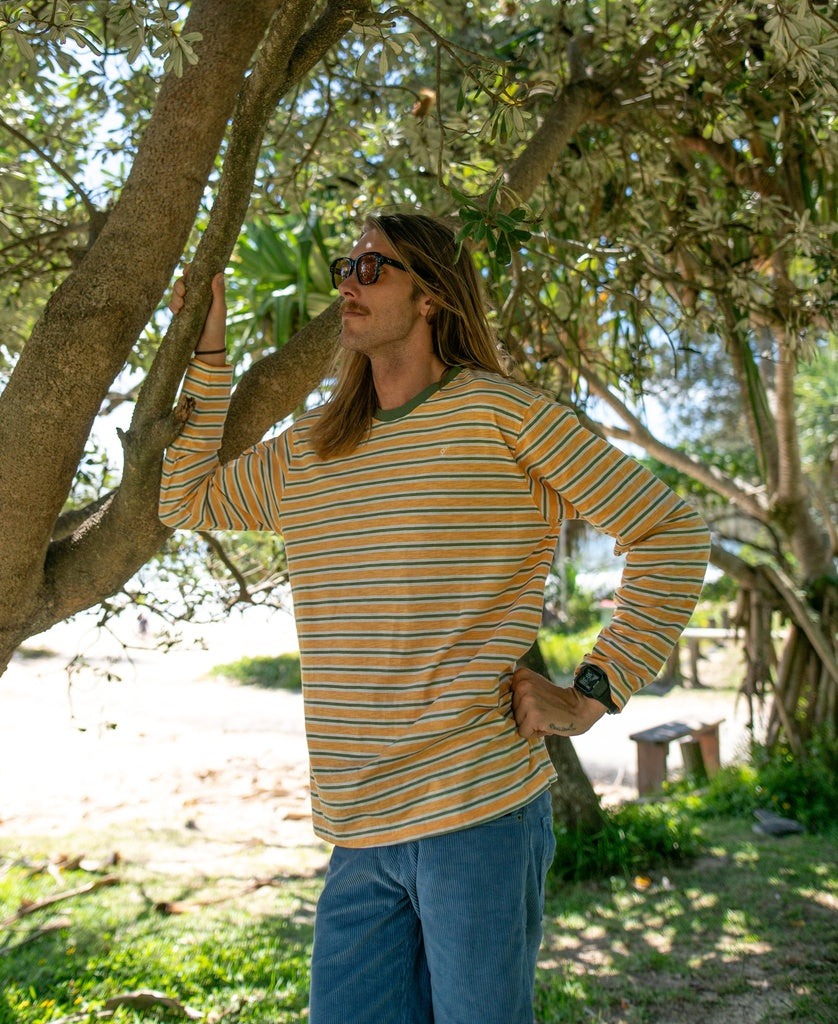 Wearing sunglasses, jeans, and a Golden Breed Rincon Stripe LS Tee in Wash Orange, a person stands outdoors under a tree, resting one arm on a branch.