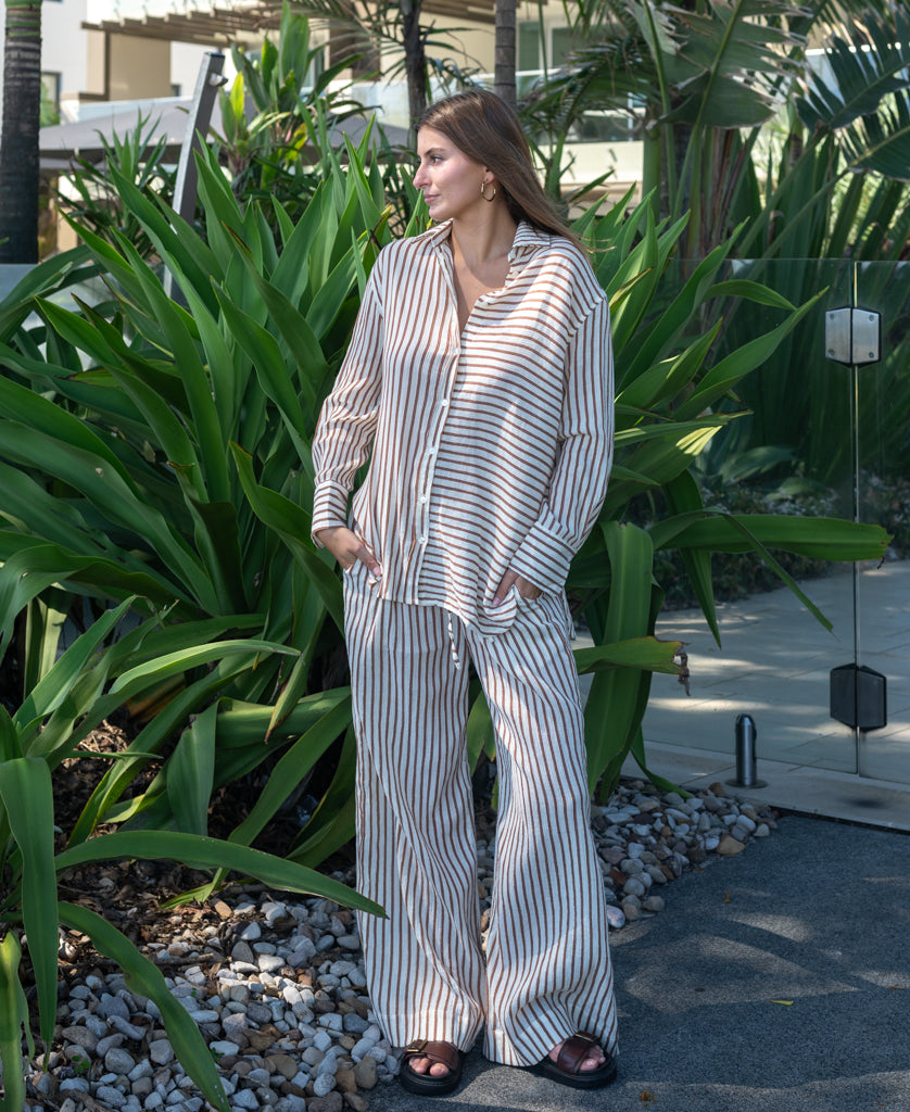 Woman standing outdoors wearing relaxed striped shirt and pants set in light brown and white, styled as Miley | Chestnut, next to greenery and glass fence.