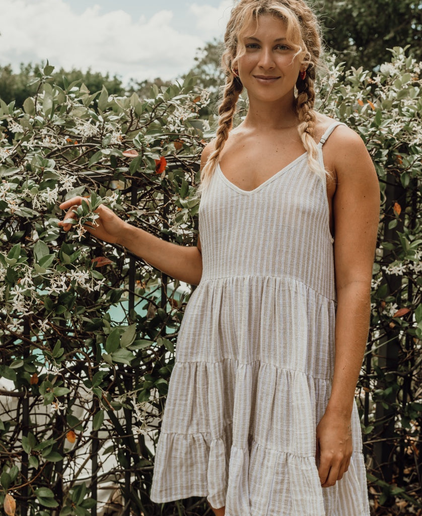 A woman with blonde braided hair wears the Golden Breed Nina Baby-doll Dress in Off White Stripe, standing before a flowering bush outdoors on a partly cloudy day.