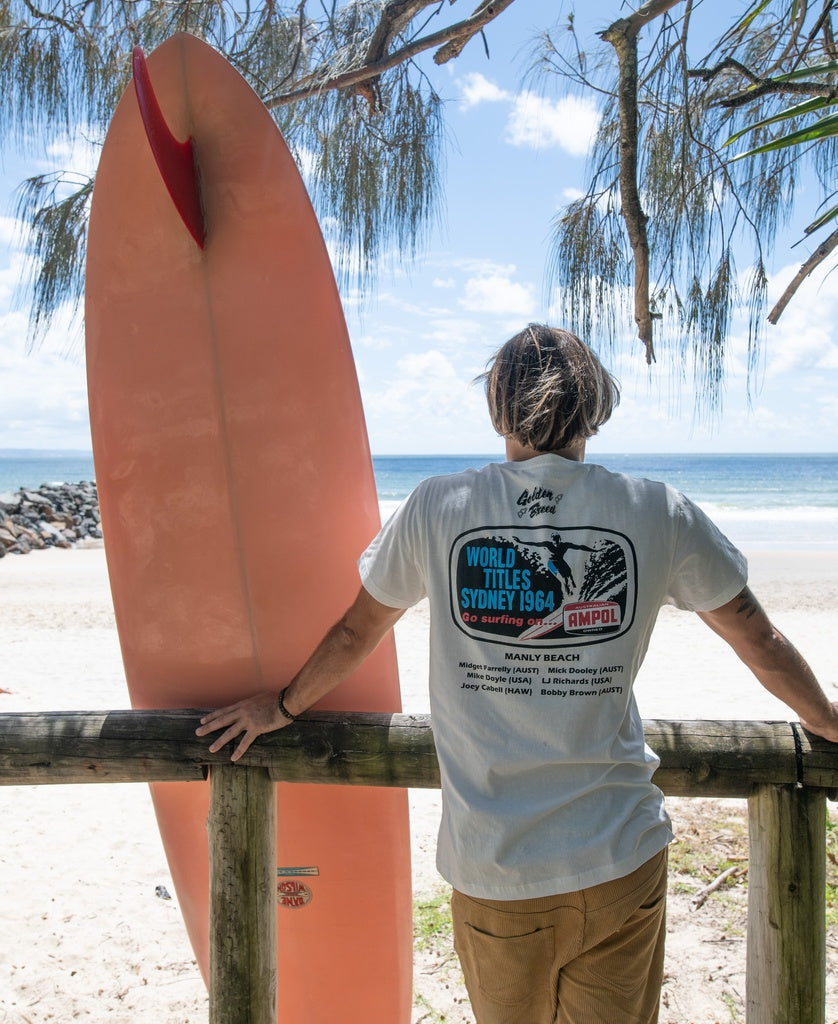 A person wearing a Golden Breed Manly Ampol Tee in Off White leans on a wooden fence by Manly Beach beside a large red surfboard, with the ocean and blue sky in the background.
