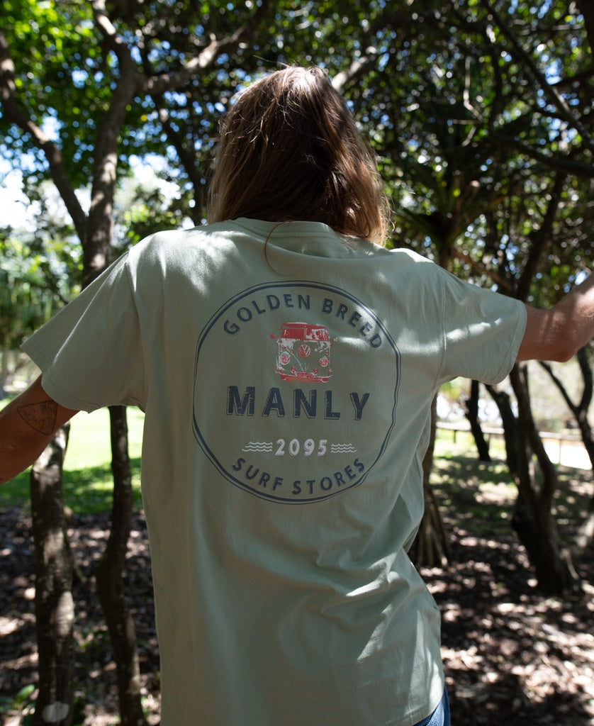 A person stands outdoors with their back to the camera, wearing the Golden Breed Manly Kombi Tee in Sage, featuring Golden Breed Manly Surf Stores 2095 and a red van graphic for authentic Aussie surf town style.
