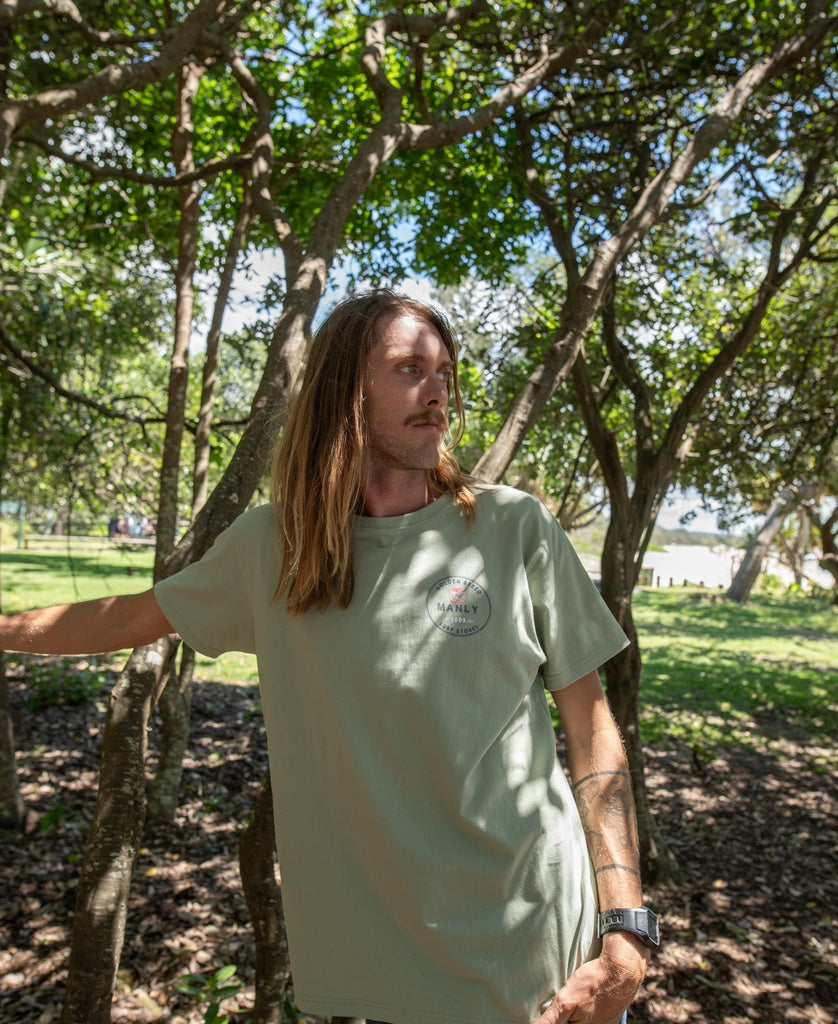 In a sunlit park, a person with long hair wears the Golden Breed Manly Kombi Tee | Sage and a watch, holding a tree branch among lush greenery.