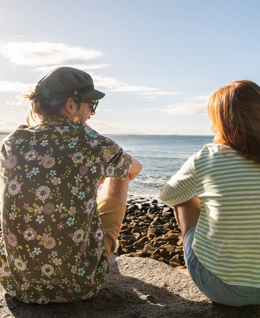 Two people sit on a rocky shore facing the ocean, one wearing the Golden Breed Happy Floral Shirt in Wash Brown and a hat, the other in a striped top. Both enjoy the sunlight.