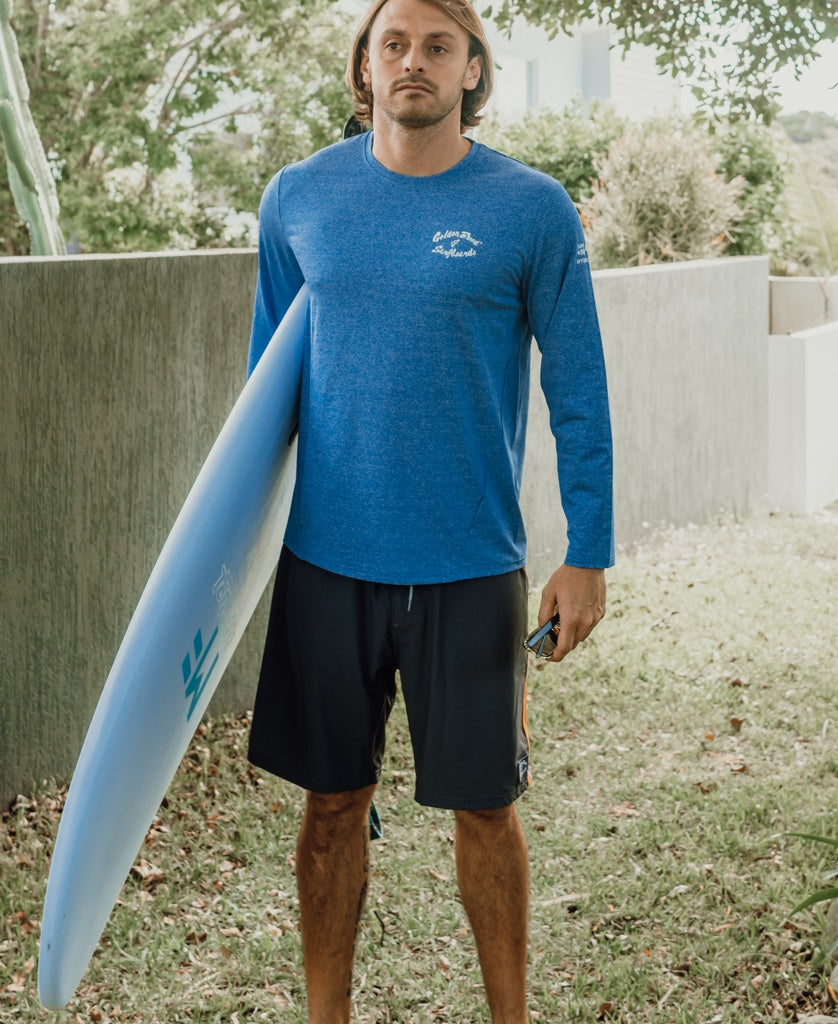 A man in a Golden Breed Pebbles L/S Rash Shirt in Blue Marle and black shorts stands on grass, holding a surfboard and phone, ready for sun protection.