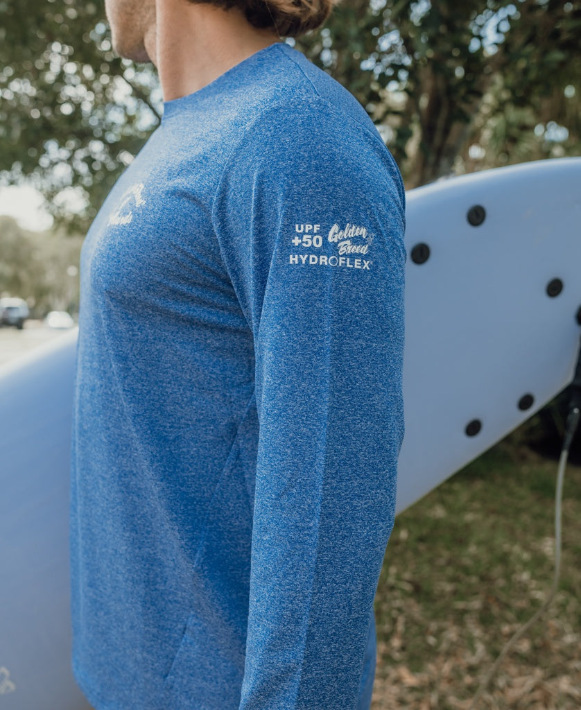 Wearing the Golden Breed Pebbles L/S Rash Shirt in Blue Marle with UPF 50+ sun protection, a person stands next to a blue surfboard outdoors.