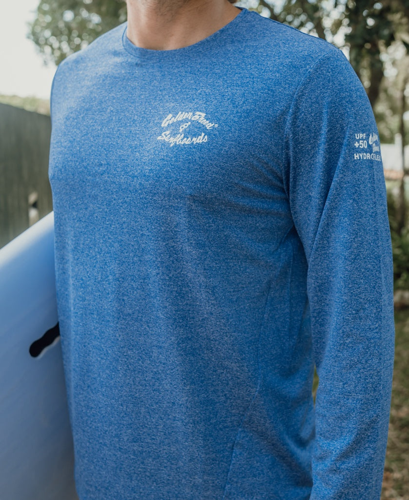 A person wearing the Golden Breed Pebbles L/S Rash Shirt in Blue Marle with UPF 50+ protection stands outdoors holding a surfboard; brand text appears on the chest and sleeve.