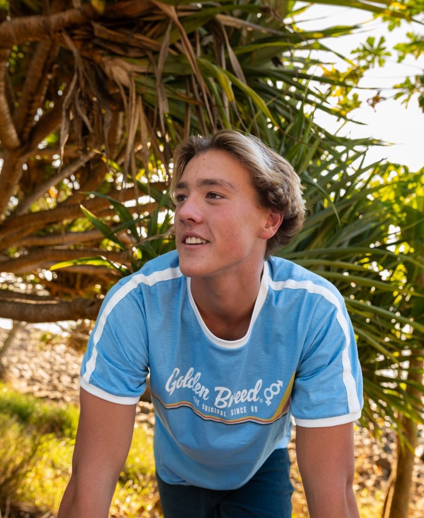 A young man smiles and leans forward outdoors near a leafy tree, sporting the cool 70โs vibes of the Golden Breed Retro Ringer Tee in Azure Blue.