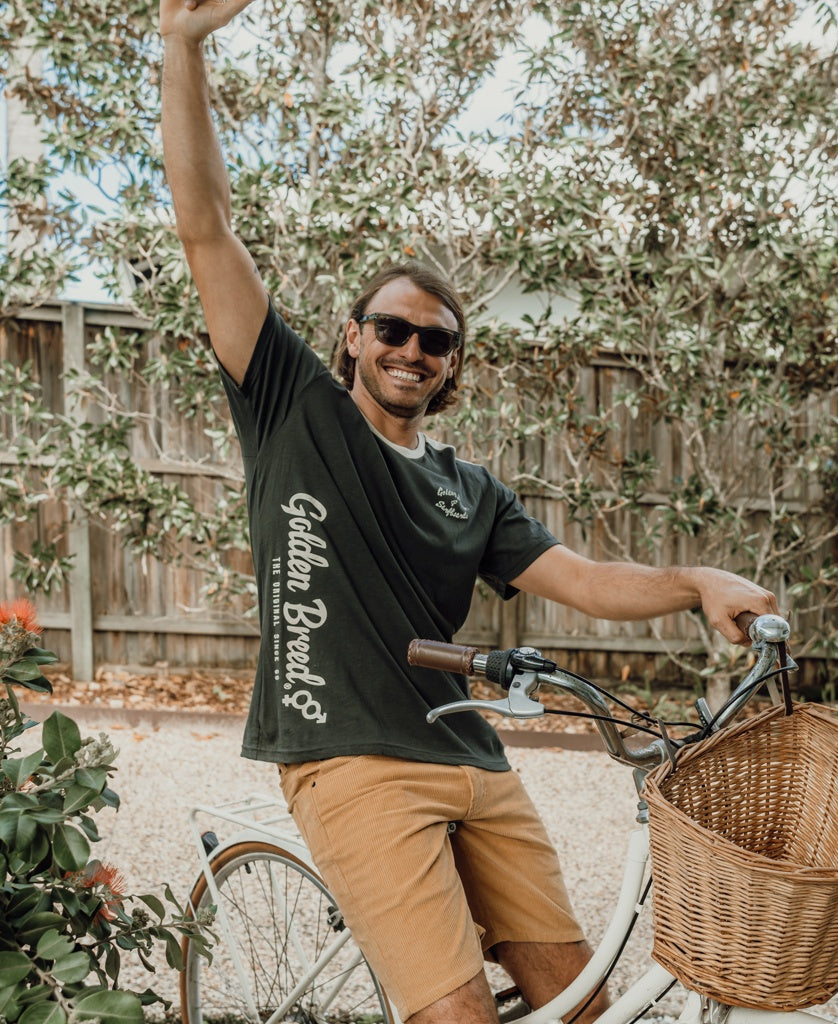 A man in sunglasses and a Golden Breed Mitch Tee | Wash Black sits on a bicycle with a wicker basket, smiling and raising one arm outdoors by a wooden fence and greenery.