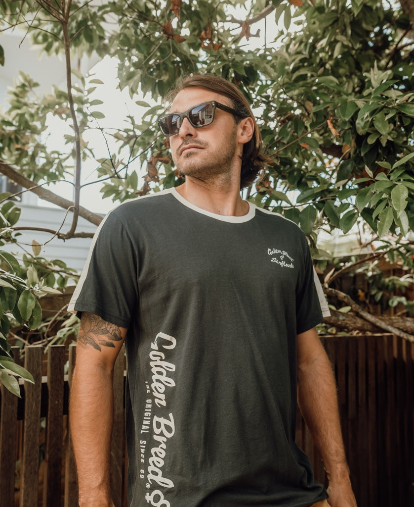 A man wearing sunglasses and a dark Golden Breed Mitch Tee in Wash Black stands outdoors in front of leafy trees and a wooden fence.