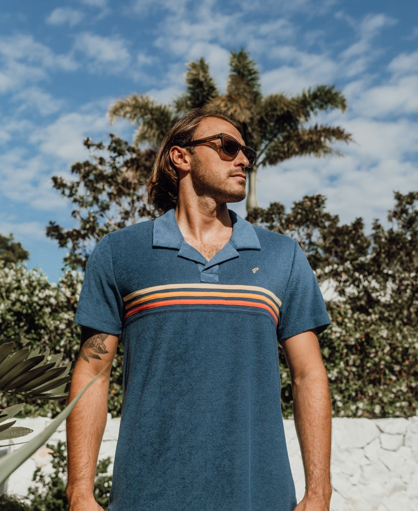 A man in sunglasses and a Golden Breed Vintage Long Island Polo | Blue, crafted from soft terry fabric, stands outdoors before trees and a palm beneath a partly cloudy sky.