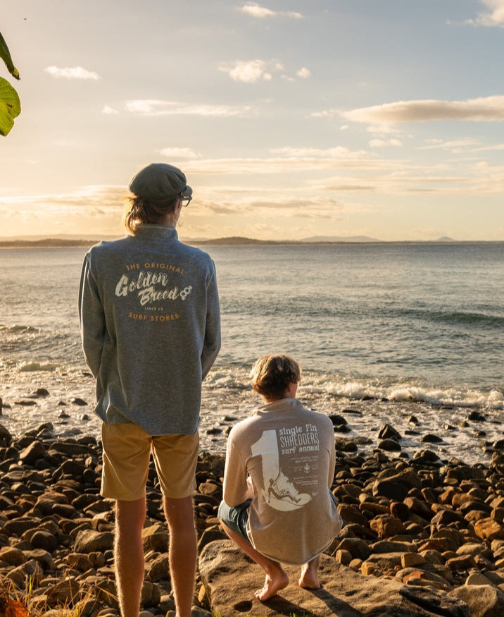 Two people stand on a rocky shoreline at sunset, both wearing Golden Breed Break 1/4 Zip Rag Top in Navy Marle with shorts. Waves crash nearby as distant landforms appear beneath a partly cloudy sky.