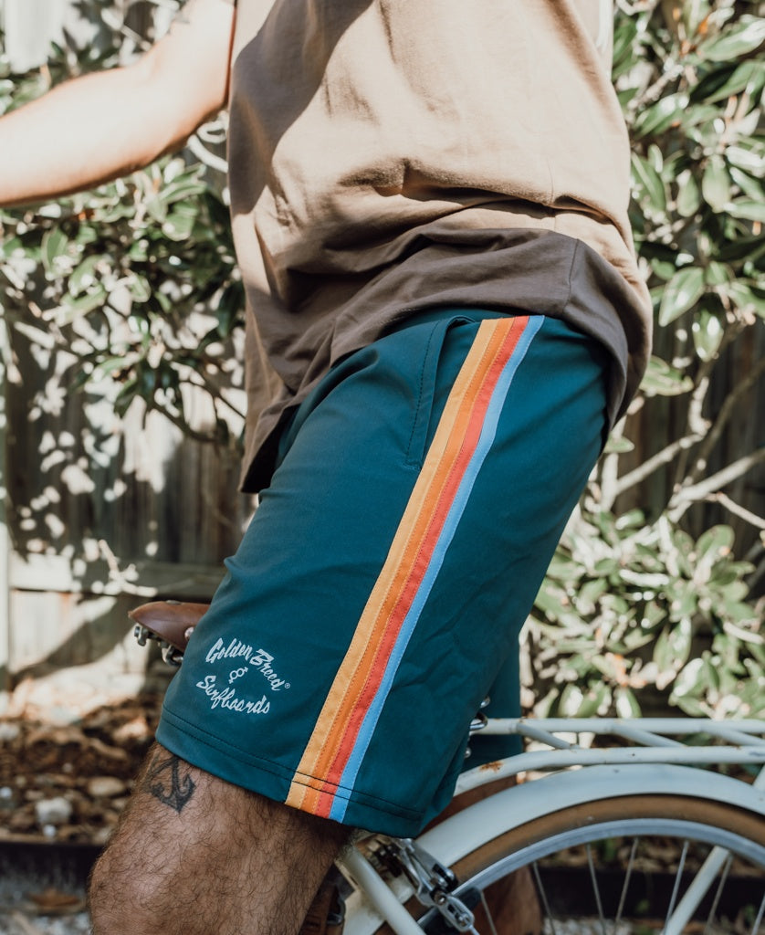 A person wearing Golden Breed Long Island Elastic Waist Boardie in Ivy Green sits outdoors on a white bicycle.