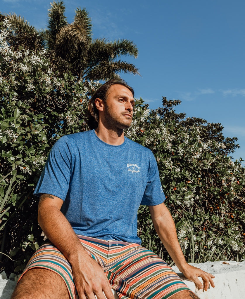 A man wears the Golden Breed Nemo S/S Rash Shirt in Blue Marle and striped shorts, sitting outside on a white surface with green foliage and a palm tree behind him beneath a clear blue sky.