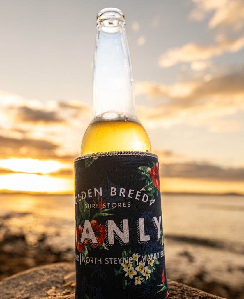 A Manly Stubby Aloha | Dark Navy by Golden Breed, featuring a floral sublimated print, stands on a rock by the water at sunset as golden sunlight shines in the background.