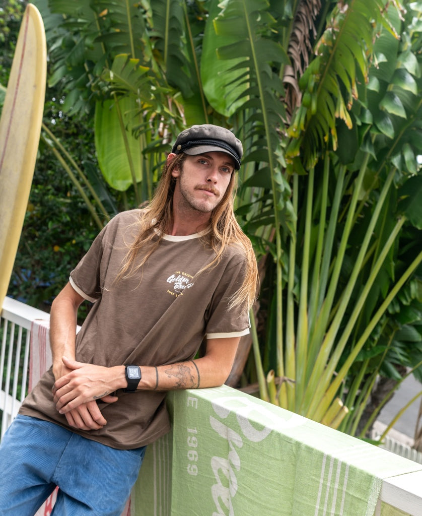 A man with long hair and a black cap stands by a railing, wearing Golden Breeds Break Ringer Tee in Wash Brown and blue pants, with large green tropical plants in the background.