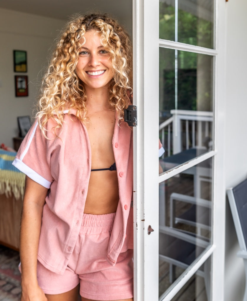 A smiling woman with curly blonde hair stands in a doorway, wearing the Golden Breed Retro Eka Shirt in Coral—handmade and dyed in Bali from soft cotton terry.