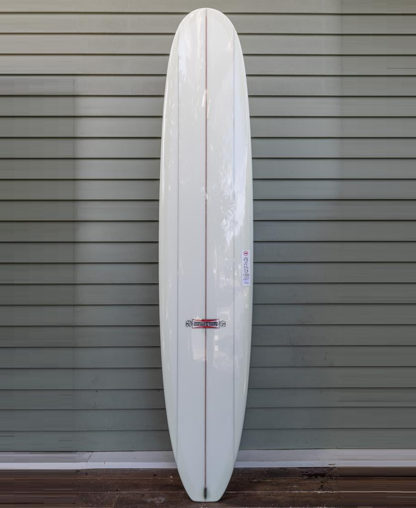 The Gordon & Smith G&S X Thomas Longboard 98, featuring a single red center stripe, stands upright against a gray paneled wall.