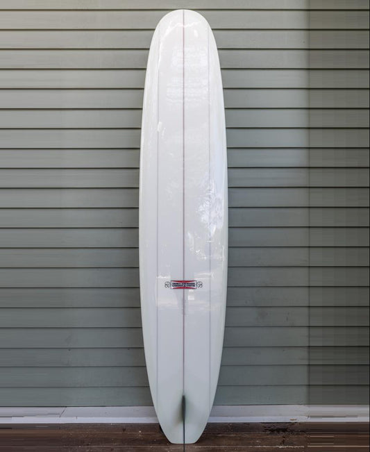 The Gordon & Smith G&S X Thomas Longboard 98, a modern performance longboard with a pin tail ideal for noseriding, stands upright against a gray horizontal siding wall.