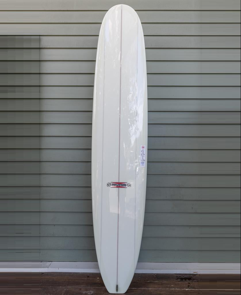 The Gordon & Smith G&S X Thomas Longboard 96, featuring a white finish with a red center stripe and designed for noseriding and modern performance, stands upright against a light gray, paneled wall.