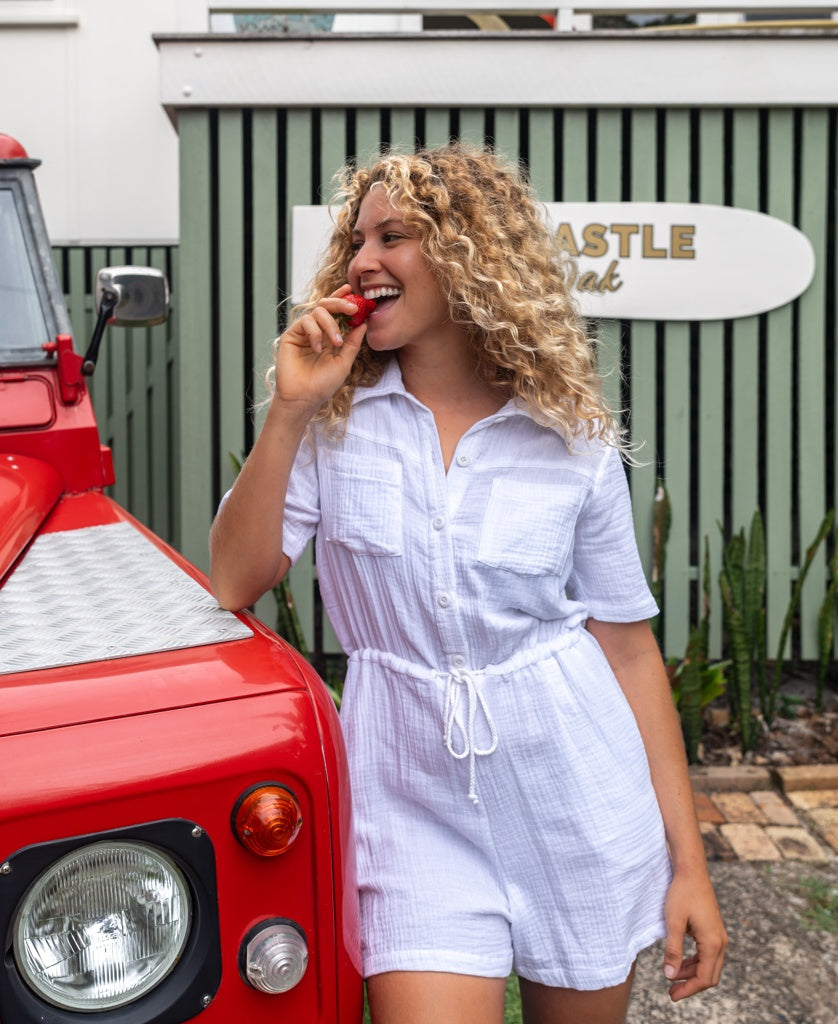 A woman in the Golden Breed Lexi Playsuit | White stands by a red vehicle, smiling with a strawberry near her mouth. A surfboard-shaped sign and green-striped fence are visible in the background.