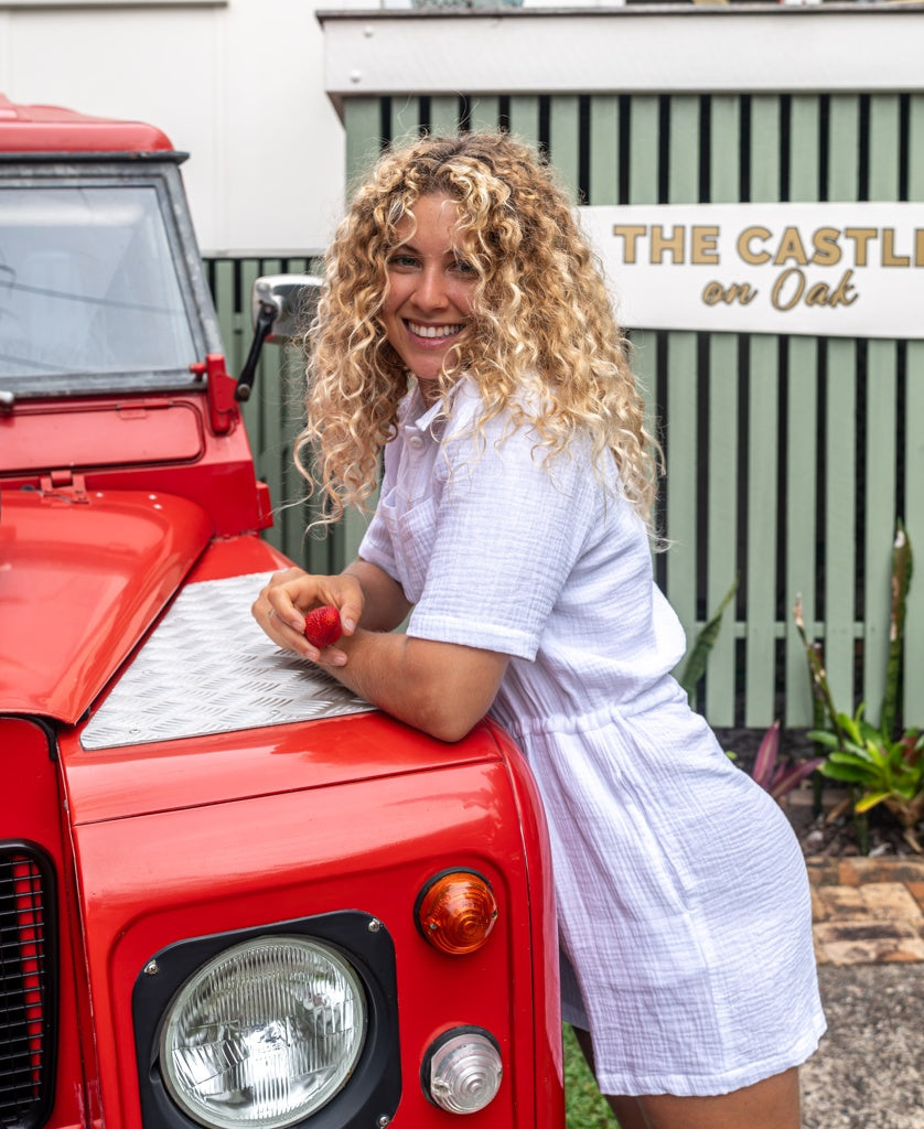 A woman with curly hair smiles while leaning on a red vehicle, wearing the Golden Breed Lexi Playsuit | White, made from a soft cotton blend. A sign reading “THE CASTLE on Oak” appears in the background.