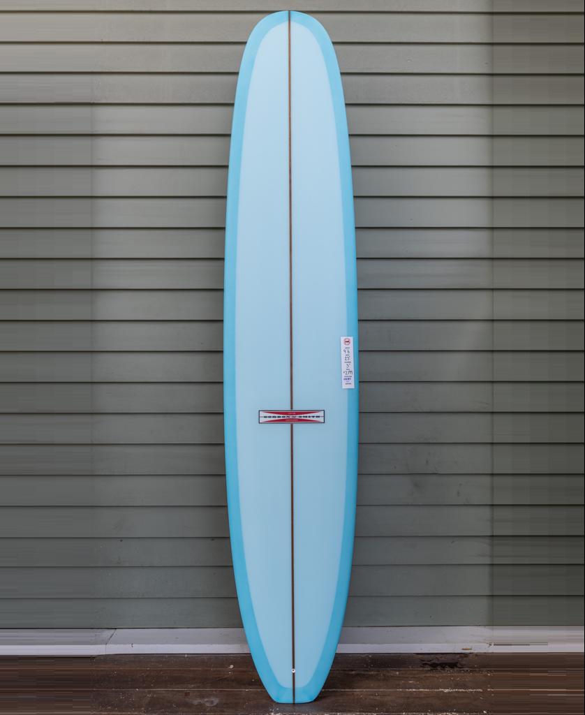 The Gordon & Smith G&S Log Baltic Sea 96 longboard, in light blue with 50/50 rails and a central wood stringer, stands upright against a gray horizontal plank wall.