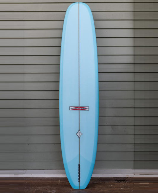 The Gordon & Smith G&S Log Baltic Sea 96, a light blue longboard surfboard with a single fin and classic 50/50 rails, stands upright against a gray horizontal-paneled wall.