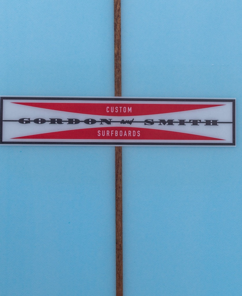 A close-up of the Gordon & Smith logo on a G&S Log Baltic Sea 96 longboard with light blue finish, 50/50 rails, and a brown stringer down the center.