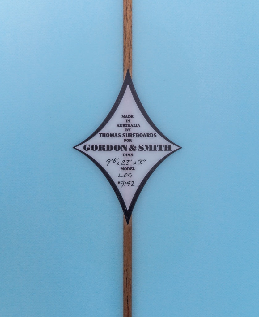Diamond-shaped label on a surfboard reads: G&S Log Baltic Sea 96 by Gordon & Smith—ideal for noseride enthusiasts. Blue background.