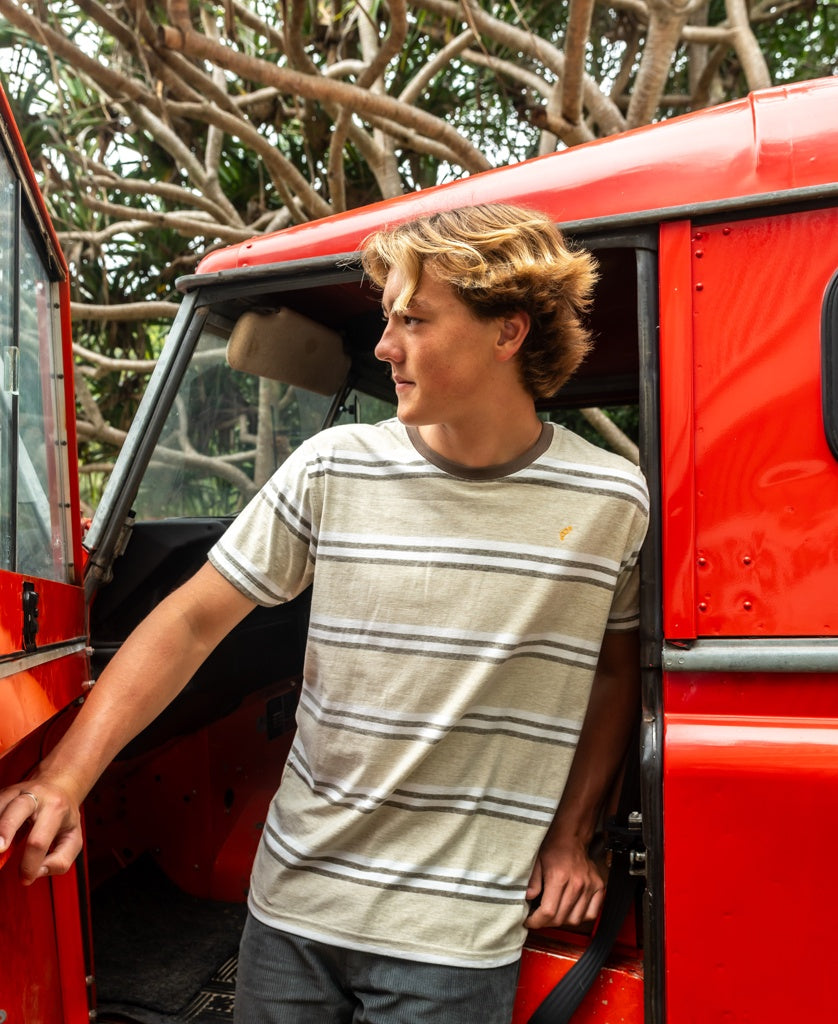 A young man wearing the Golden Breed Bay Stripe Tee in Mocha stands beside the open door of a red car, looking to the side with twisted trees in the background.