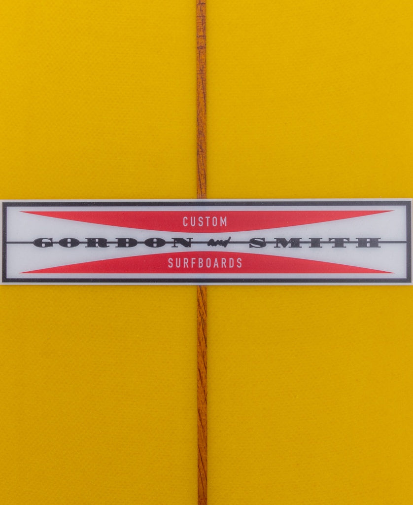 Close-up of the G&S Log Gold Fusion 94 by Gordon & Smith, showing a yellow board with a centered wooden stringer, classic rails, and a label with red and black text.