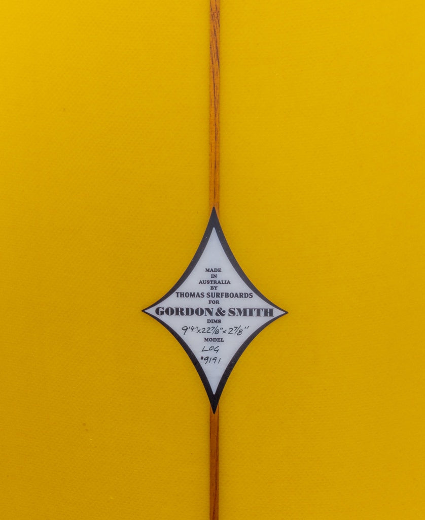 Close-up of a diamond-shaped label on the yellow G&S Log Gold Fusion 94 by Gordon & Smith, showcasing manufacturer info and dimensions—ideal for surfers craving a classic longboard feel with smooth noseride performance.