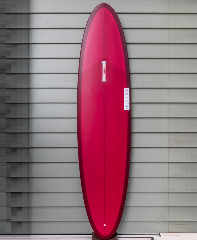 The Gordon & Smith G&S Mid length Cardinal 76, a red surfboard with a pointed nose and rounded tail, stands upright against a gray horizontal paneled wall.