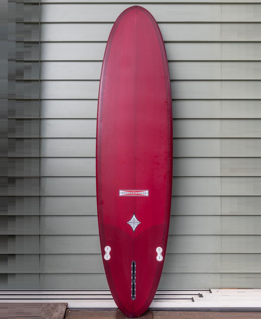 The Gordon & Smith G&S Mid length Cardinal 76 surfboard, featuring a rounded nose and fin boxes, stands upright against a gray horizontal panel wall—a versatile red board perfect for any quiver.