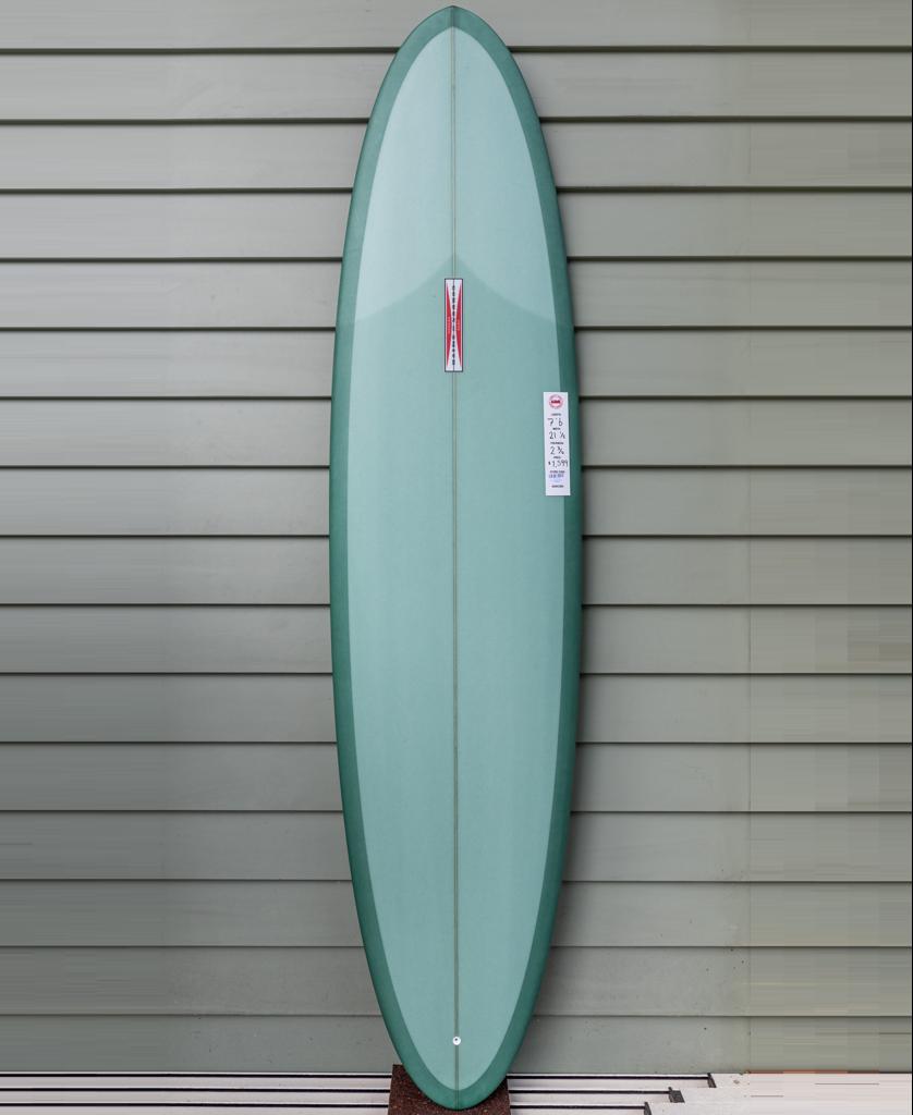 A Gordon & Smith G&S Mid length Malachite Green 76 surfboard with a pointed nose and rounded tail stands upright against a gray horizontal paneled wall.