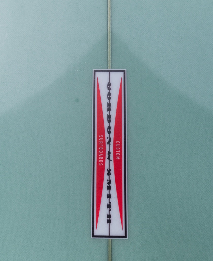 A rectangular logo on the G&S Mid length Malachite Green 76 surfboard by Gordon & Smith features Gordon & Smith Custom Surfboards in black and red text on a white background with red accents.