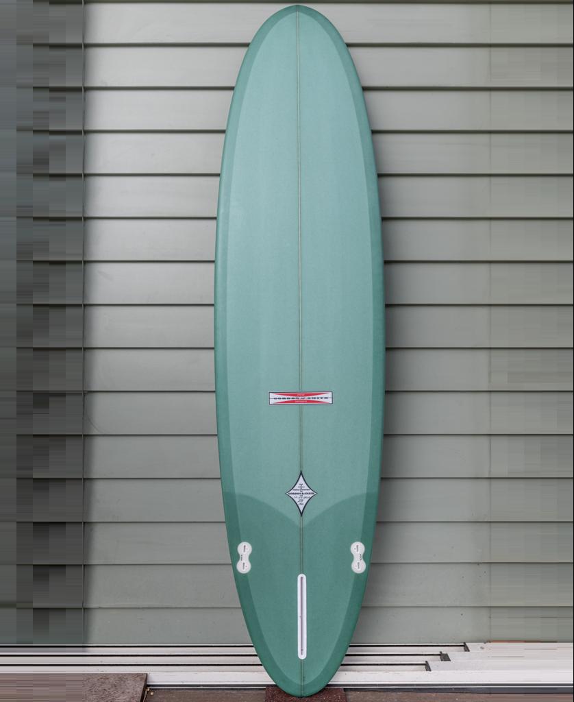 A Gordon & Smith G&S Mid length Malachite Green 76 surfboard with a pointed nose and rounded tail stands upright against a gray paneled wall.