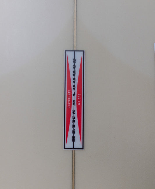 A rectangular sign with a red and white geometric design reads Gordon & Smith G&S Long Twin Sahara Sun 68, mounted on a light-colored wall.