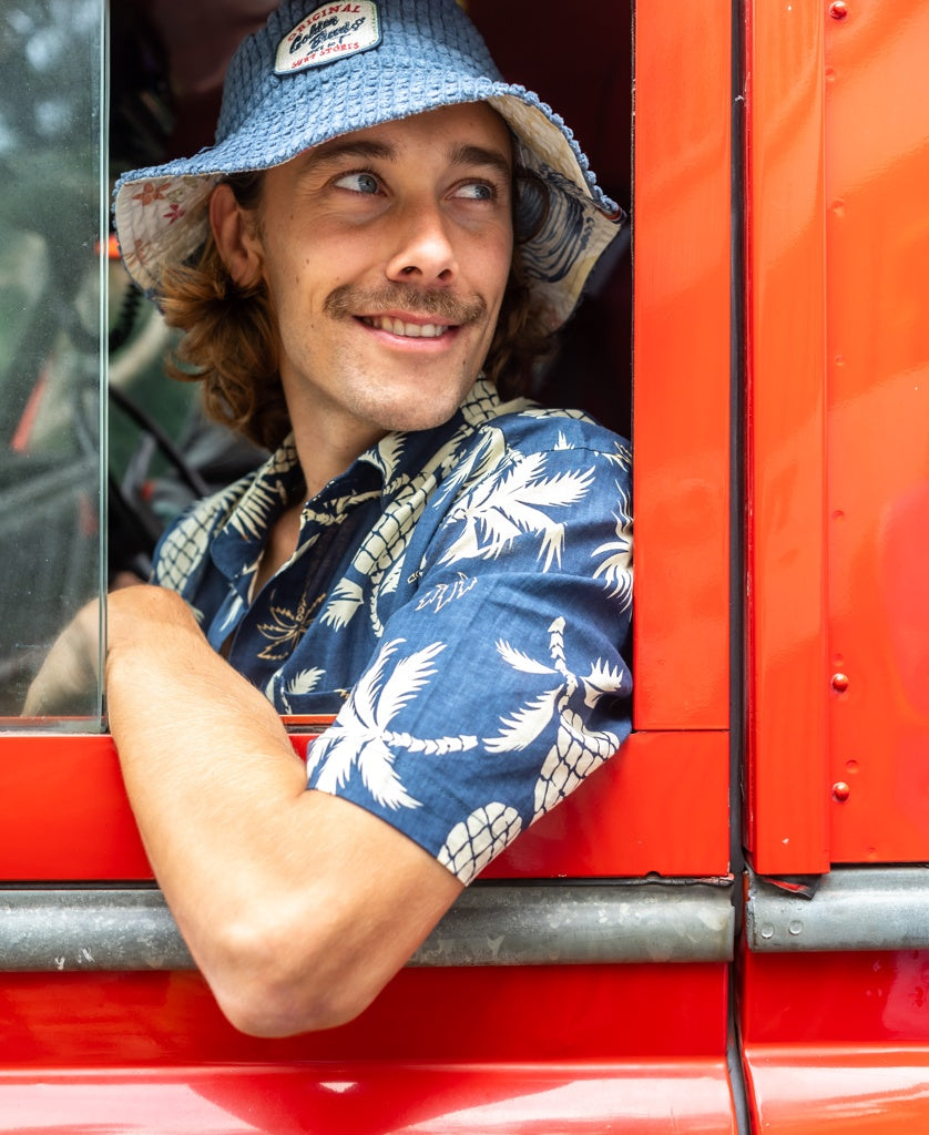 A man wearing a blue patterned shirt and the Golden Breed El Noggino Cord Bucket Hat in Denim smiles from the drivers seat of a red vehicle, his arm resting on the window frame.