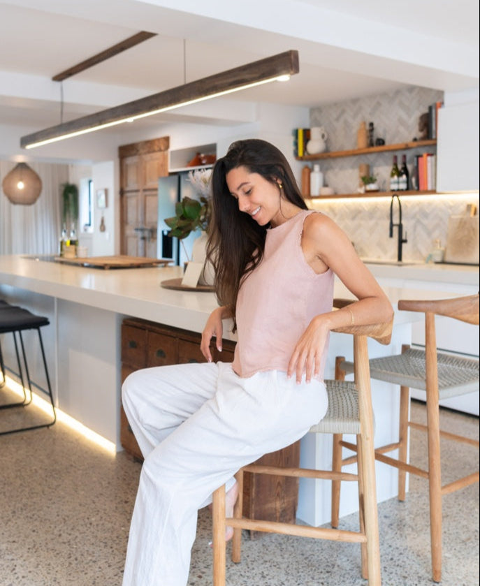 A woman wears the Lazy Beach Angelina | Blush Linen sleeveless top with frayed trim and white pants, sitting on a wooden chair in a modern kitchen with open shelves and a large island.