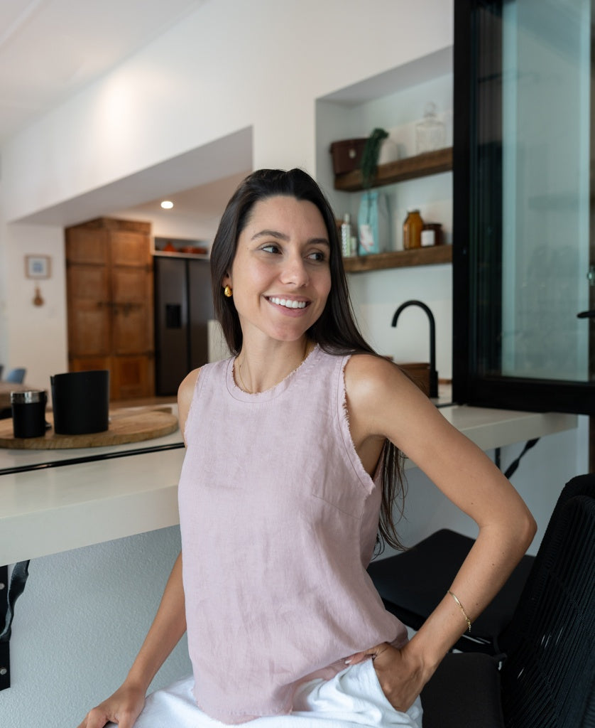 A woman with long dark hair wears the Lazy Beach Angelina blush linen sleeveless top and white frayed pants, smiling in a modern kitchen with open shelving and a black faucet.