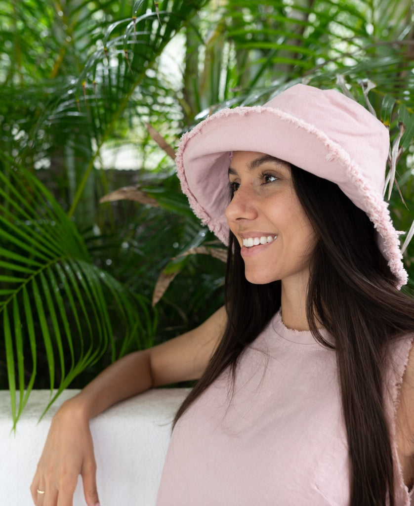 A woman wears the Lazy Beach Halo Bucket Hat in Blush, standing by a white wall and smiling with green tropical plants in the background.