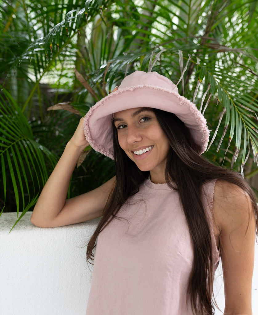 A woman in a light pink sleeveless top and the Lazy Beach Halo Bucket Hat in Blush stands by a white wall, smiling, with green foliage behind her.